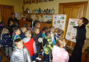 Grupa dzieci z panią Ewą spoglądają na panią w czarnym stroju, która prawą ręką wskazuje na planszę o produkcji mleka i sera